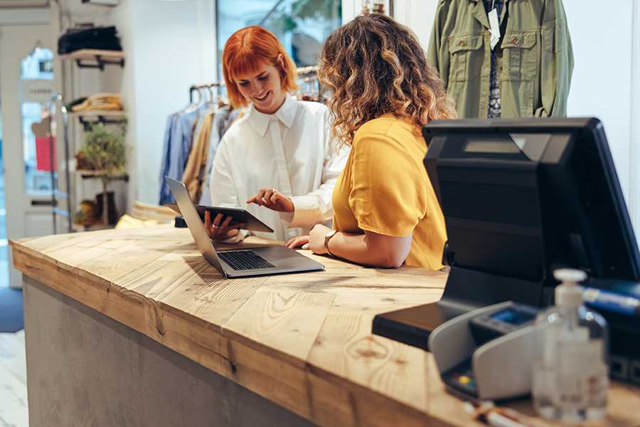 Clothing store workers standing at checkout counter using digital tablet. Two fashion shop partners looking at online orders on a tablet computer in store.